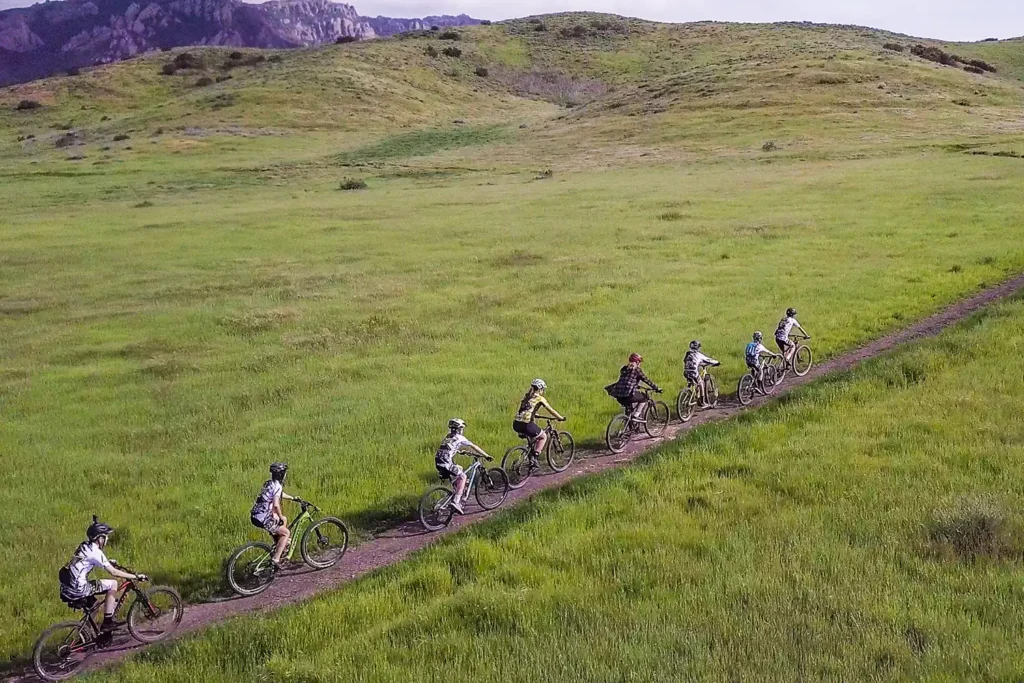 A group of people ride mountain bikes single file along a trail in a grassy field.