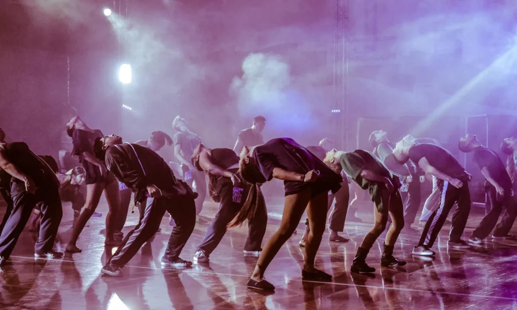 Multiple dancers leaning backwards in a synchonized dance on a stage with purple lighting and fog.