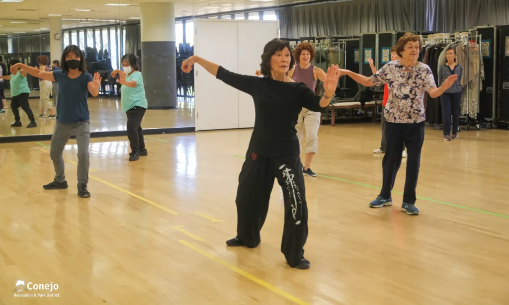 Older people with their arms outstretched practicing tai-chi inside a studio with mirrored walls.