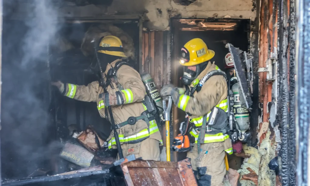 Firefighters in full gear navigating through a burnt building with smoke.